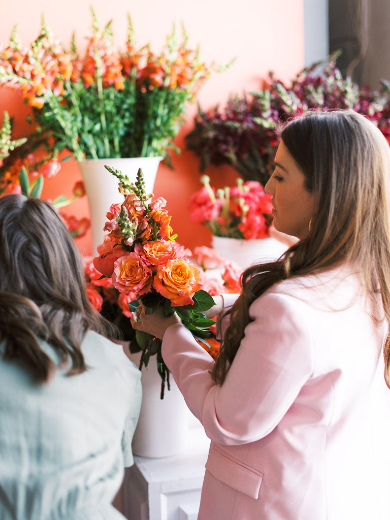 Two women, with their backs facing the camera, pick flowers out of large white vases and buckets to add to the bouquets they are arranging at Flower Hoopla, a floral workshop in Alexandria, Virginia.