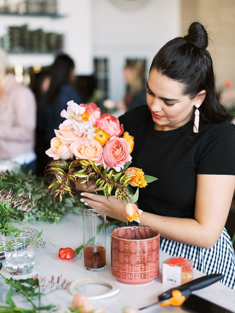A woman with dark brown hair in a top-knot, who is wearing bright pink lipstick, dangly earrings, and a black, fitted t-shirt, works on designing her floral arrangement at a floral workshop. There are various flowers, tools, and greenery on the table at her work station. Taken by Maryland and Virginia commercial photographer Kim Branagan.