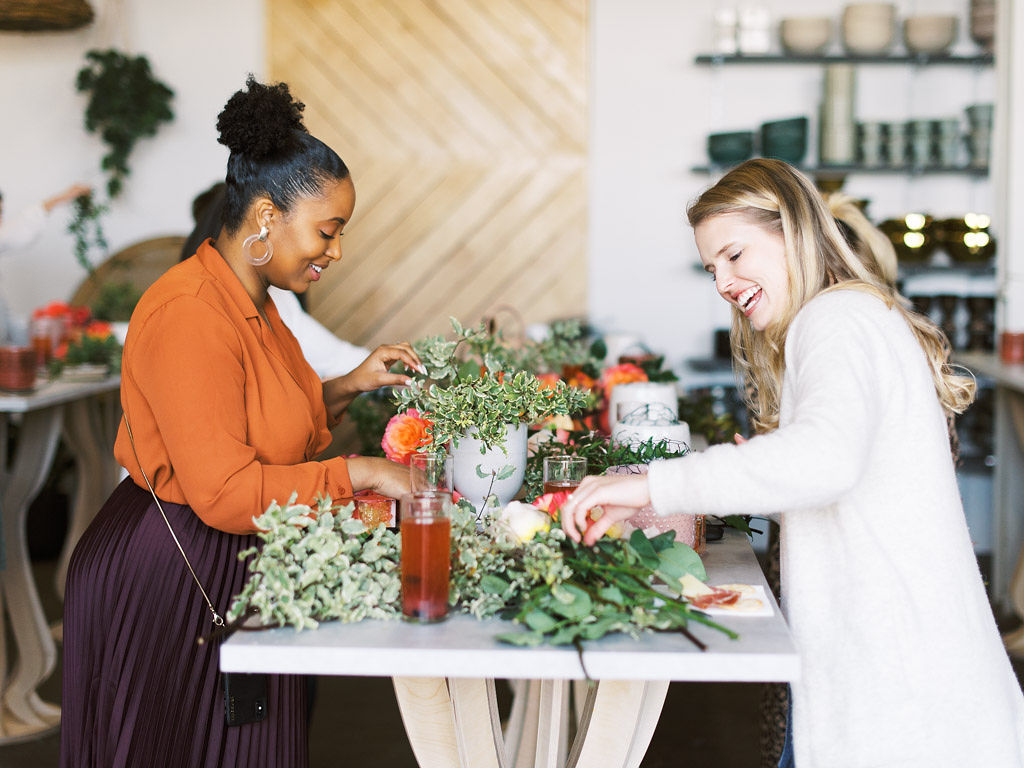 Two women laugh as they work on designing their flower arrangements at a floral workshop. There are various flowers, tools, and greenery on the table at her work station. Taken by Maryland and Virginia commercial photographer Kim Branagan.