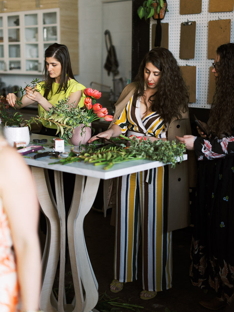 Two women stand side by side at a tall wooden table serving as their work station at a floral workshop. The women have lots of greenery in front of them and are adding more flowers and stems to the vases full of flowers in front of them.