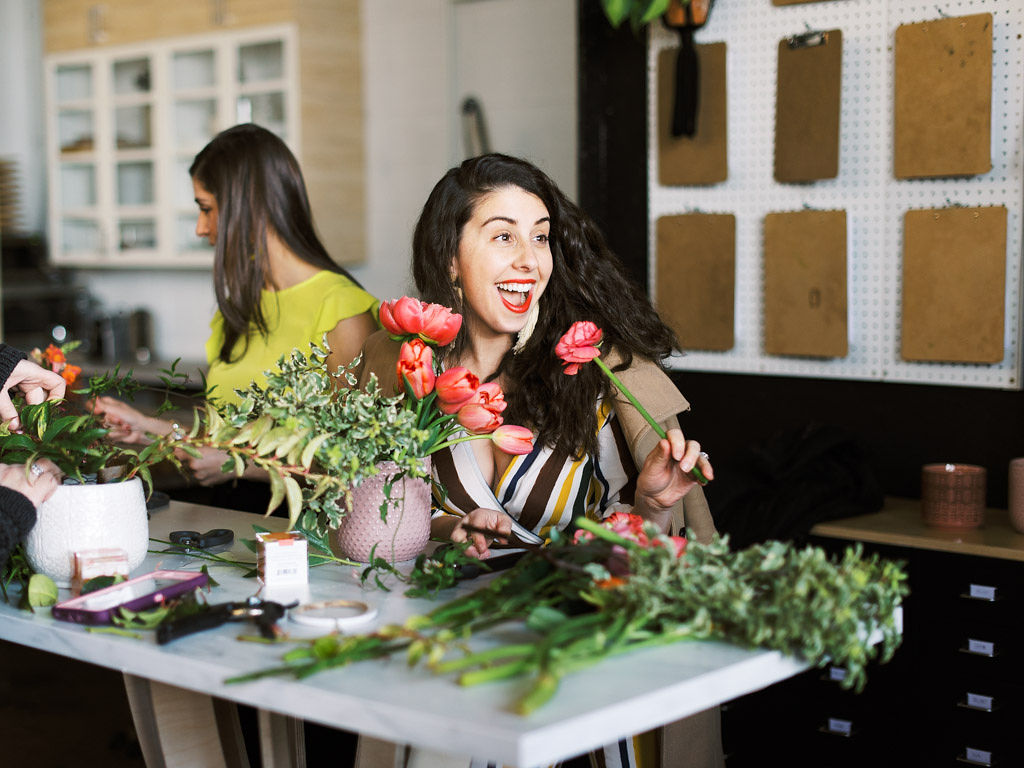 A woman smiles widely with her mouth open and holds a bright pink peony up to her mouth. She is standing at her floral design work station, working on putting together a bouquet of pink peonies and tulips with lush greenery. Greenery, flowers, and tools lay on the table in front of her. Taken by D.C. commercial photographer Kim Branagan.
