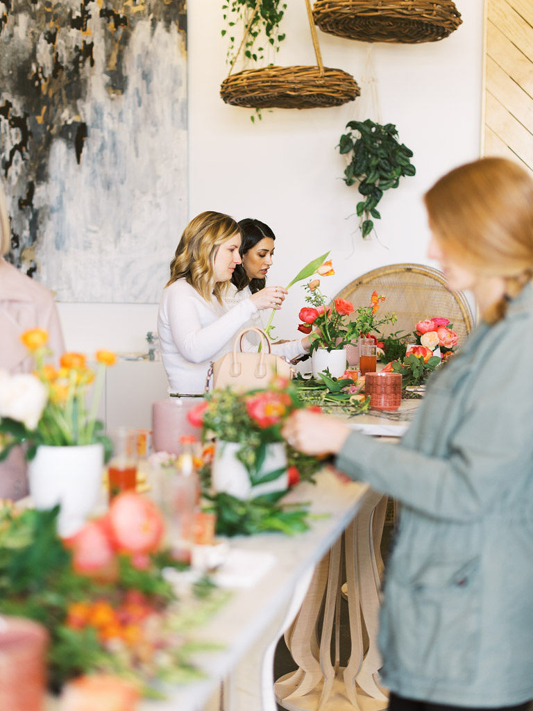 A long, tall wooden table serves a work station for several women attending a floral design workshop at Sweet Root Village in Alexandria, VA. There are vases with floral bouquets, loose flowers and greenery, beverage cups, and tools all over the table.