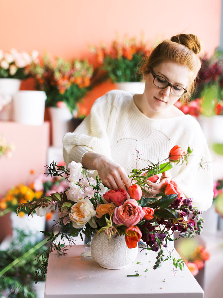 A florist places her hands on a foral bouquet as she works to design it. The pink, white and red garden roses sit in a white ceramic vase.