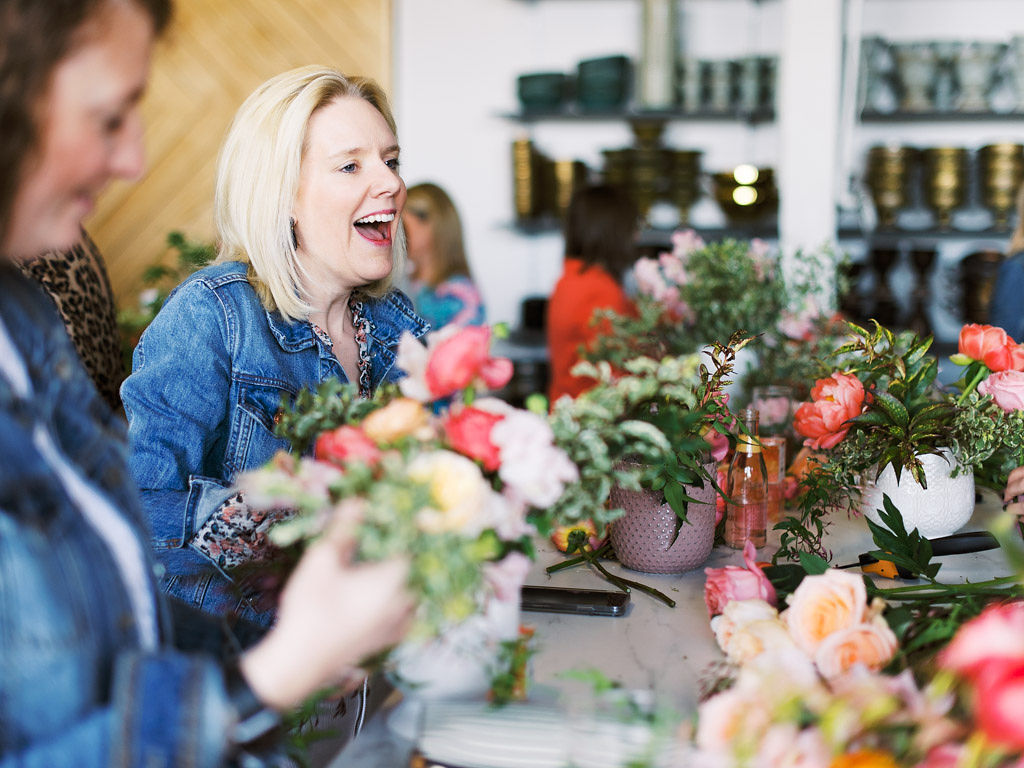 A woman smiles and laughs as she works on creating a flower arrangement at her work table. Several vases of bouquets are in front of her and other women are working on their bouquets in the background.