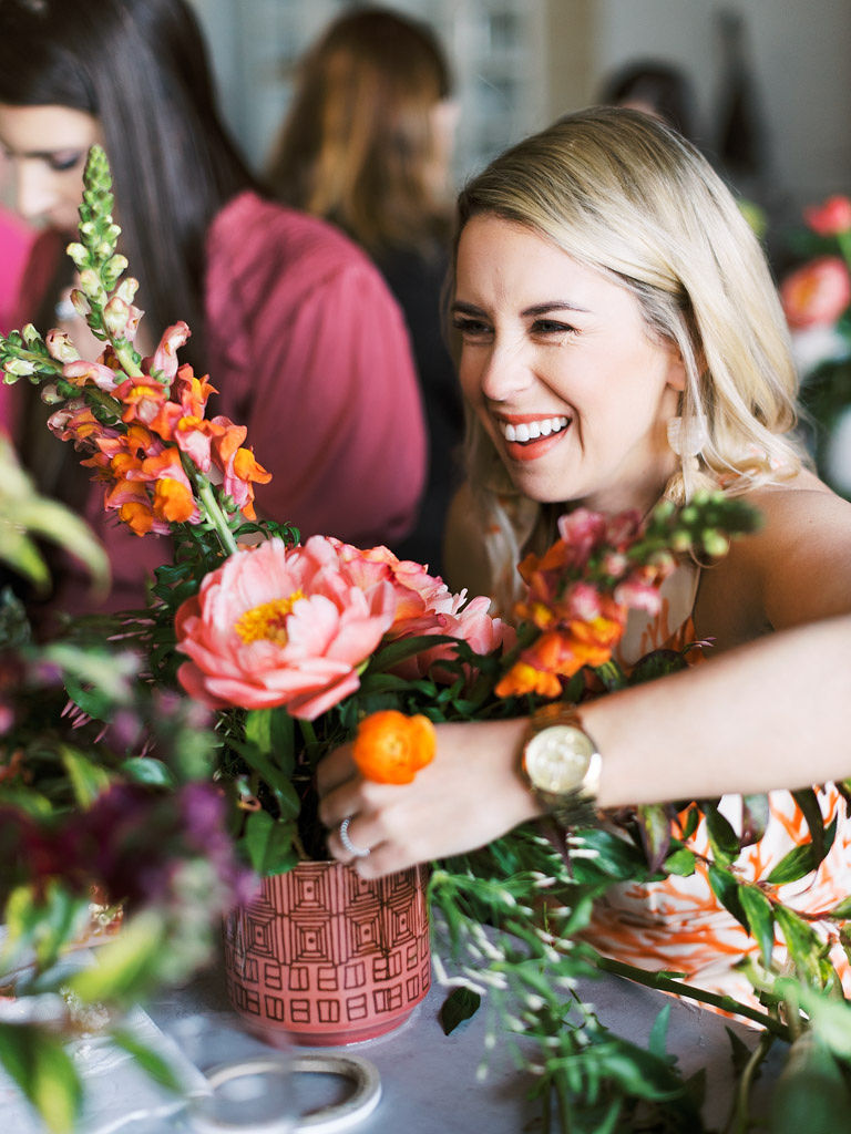 A blonde-haired woman smiles as she places a small, orange flower in a maroon vase with black geometric patterns on it. The vase is filled with other bright orange, pink, and yellow flowers and long-stemmed greenery. Taken by Virginia commercial photographer Kim Branagan.