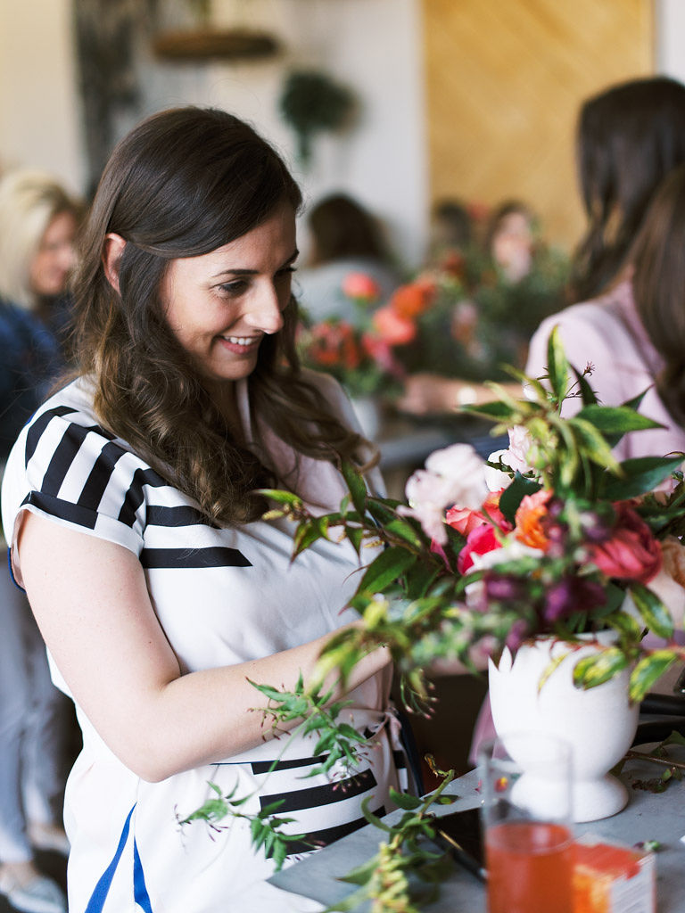 A woman wearing a white dress with black striped geometric shapes on the shoulder and abdomen of the garment arranges flowers in her white, orange, and pink floral bouquet at the Flower Hoopla workshop at Sweet Root Village.