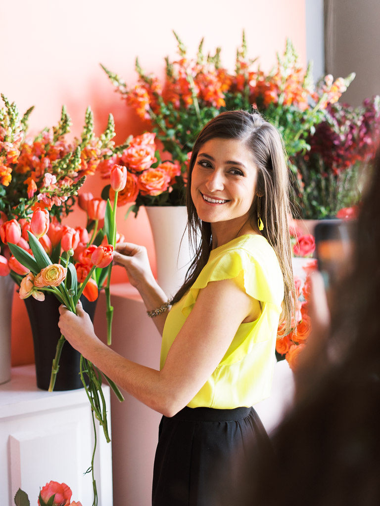 A woman picks out pink tulips from a bucket full of tulips at Sweet Root Village. The left side of her body is facing the camera, and she is looking over her shoulder and smiling.