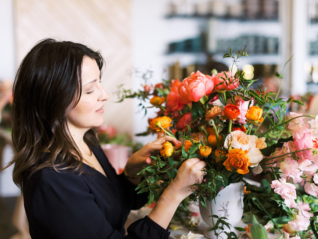 A woman wearing a black blouse arranges flowers and greenery in a lush bouquet of pink, red, and orange flowers. Taken by D.C. wedding and commercial photographer Kim Branagan.