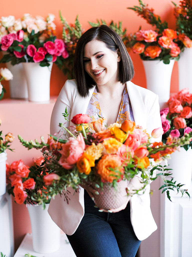 A woman with should-length brown hair wearing a white blazer and jeans holds her floral arrangement made of greenery and flowers that are various shades of pink and orange. The floral arrangement is in a light pink vase. Photo by Kim Branagan, a greater DMV area commercial and wedding photographer.