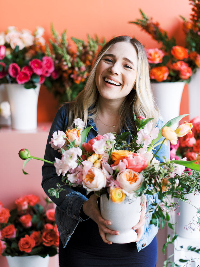 A woman with long blonde hair smiles as she holds the pink, orange, and green bouquet she design at the Flower Hoopla workshop in Alexandria, Virginia.