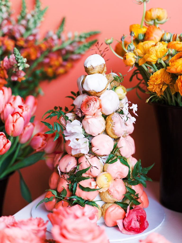 A tower of donut holes covered in various shades of pink frosting with springs of greenery and flower buds stuck into the arrangement. The pastries are on a table surrounded by bouquets of pink, red, and orange flowers. Taken at Sweet Root Village by D.C., Virginia, and Maryland commercial photographer Kim Branagan.