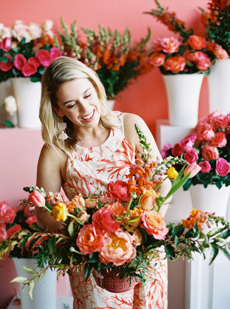 A woman wearing a cream -colored dress with a coral design on it smiles and looks down at the large bouquet of pink, orange, and yellow flowers she designed. Lots of pink roses and peonies are behind her. Photo taken by D.C. an Virginia commercial photographer Kim Branagan.