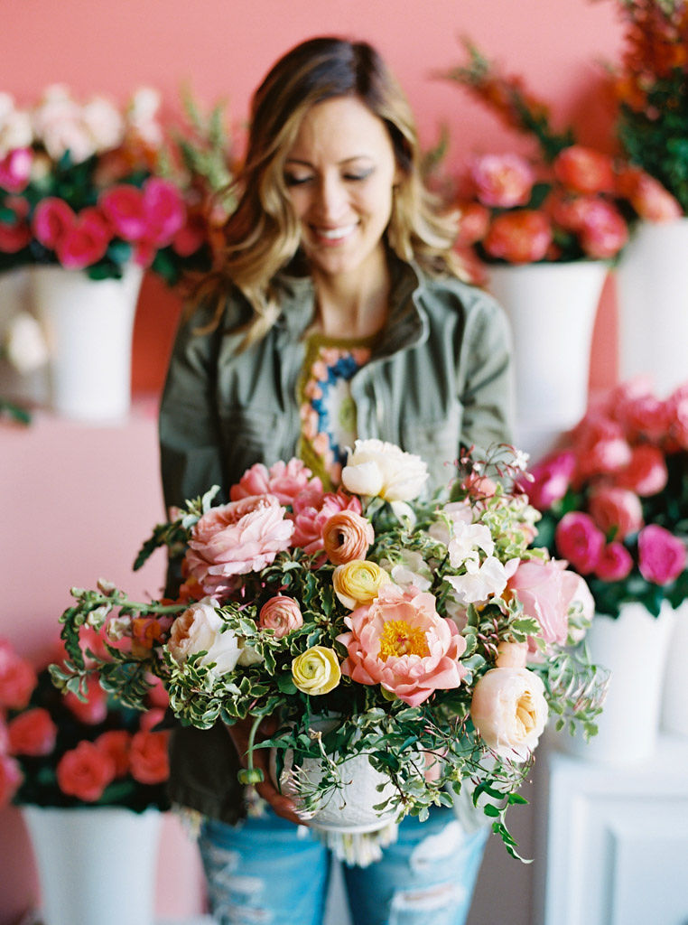 A woman wearing a gray canvas jacket stands in front of a pink wall and lots of bouquets of pink flowers. She is holding a floral bouquet comprised of white, yellow, and pink flowers with lots of greenery.