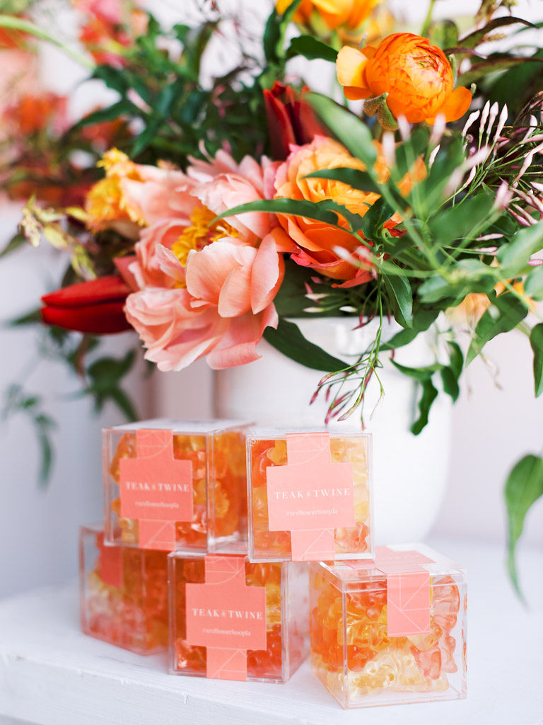 Small, clear boxes of Teak & Twine peach gummy bears, with a colorful bouquet of orange, pink, and red flowers with lots of greenery in the background. Taken at the Sweet Root Village studio in Alexandria, VA.