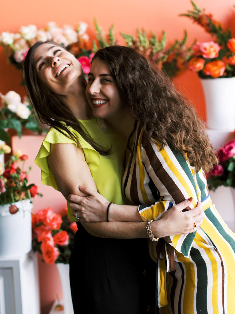 Two women laugh and hug standing in front of a coral pink wall with lots of bright pink and white flowers in the background. Taken by Virginia wedding and commercial photographer Kim Branagan at Sweet Root Village, Alexandria, VA.