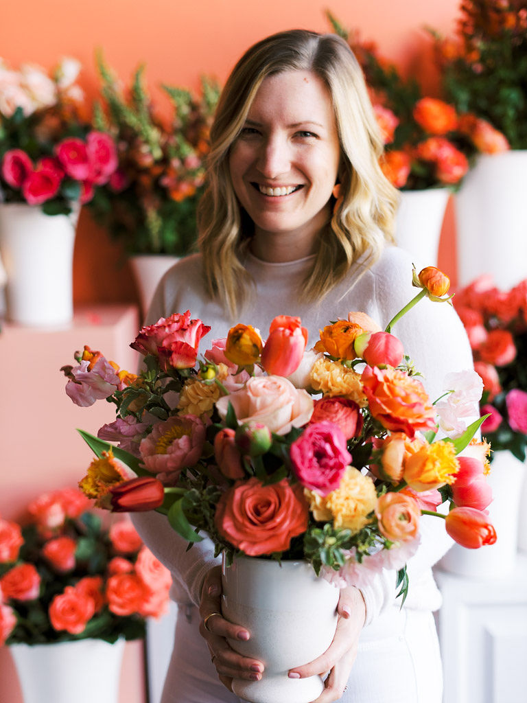 A women wearing a white, mock-neck sweater and white jeans holds a colorful bouquet of dark pink, light pink, burnt orange, and dark yellow flowers in a white vase.