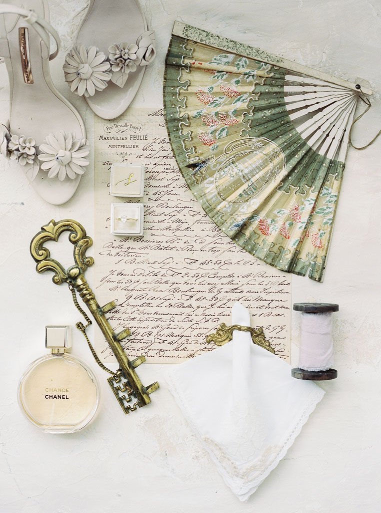 Close up shot of trinkets from a wedding day: the bride's shoes, handwritten vows, a paper fan, the wedding rings, Chanel perfume, and an old-fashioned key.