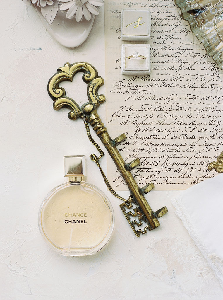 Close up shot of trinkets from a wedding day: handwritten wedding vows on paper, the wedding rings, Chanel perfume, and an old-fashioned key. Taken at Antrim 1844 in Maryland, by Maryland wedding photographer Kim Branagan.