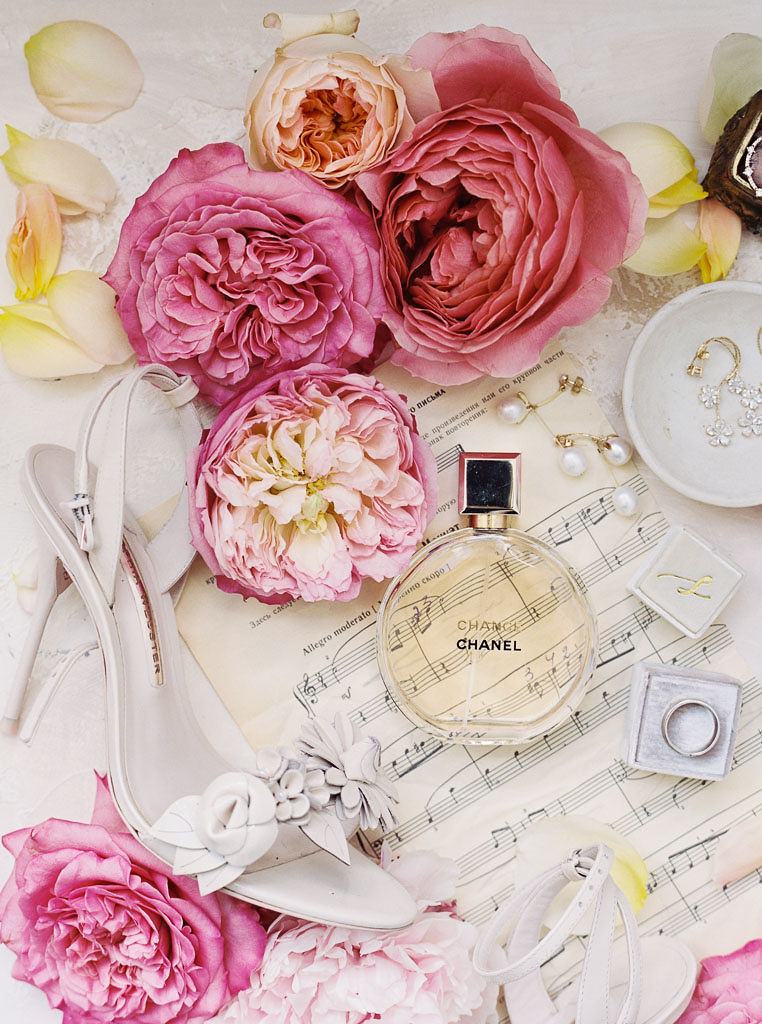 Close up shot of trinkets from a wedding day: sheet music, bright pink flowers, the wedding rings, the bride's white high heels, and Chanel perfume. Taken at Antrim 1844 in Maryland, by Maryland wedding photographer Kim Branagan.