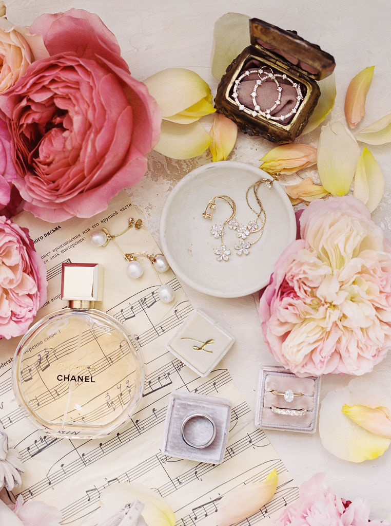 Close up shot of various objects: Pink flower blossoms, gold jewelry, Chanel perfume, sheet music, and scattered light yellow flower petals.