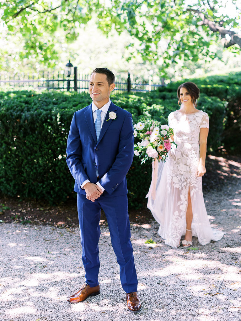 A couple doing their first look on their wedding day. The bride is standing behind the groom; he has his back toward her. He is wearing a blue suit and is smiling. The bride is wearing a lace wedding gown and holding a large pink and white bouquet. They are standing in a garden at Antrim 1844 in Taneytown, Maryland.