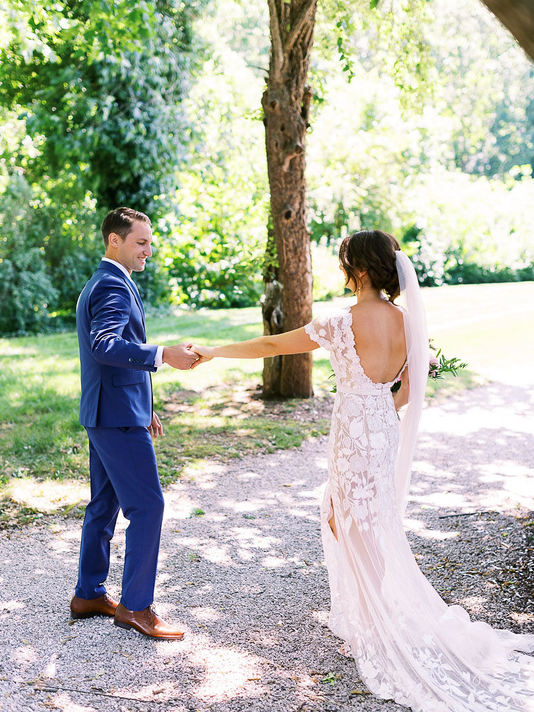 A couple holding hands and walking during their first look on their wedding day. They have their backs to the camera, and they are holding hands, looking at each other. They are on a gravel path in the forest.