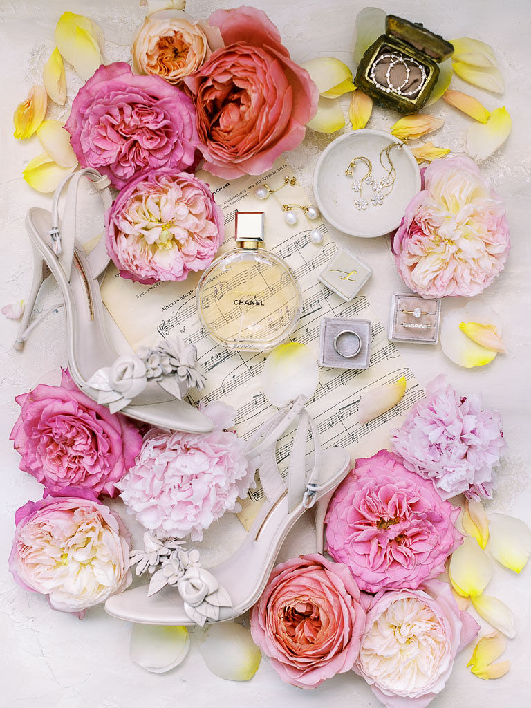 Rectangular shot of various objects arranged on a white background. The objects are: Pink flower blossoms, gold jewelry, Chanel perfume, sheet music, and scattered light yellow flower petals.