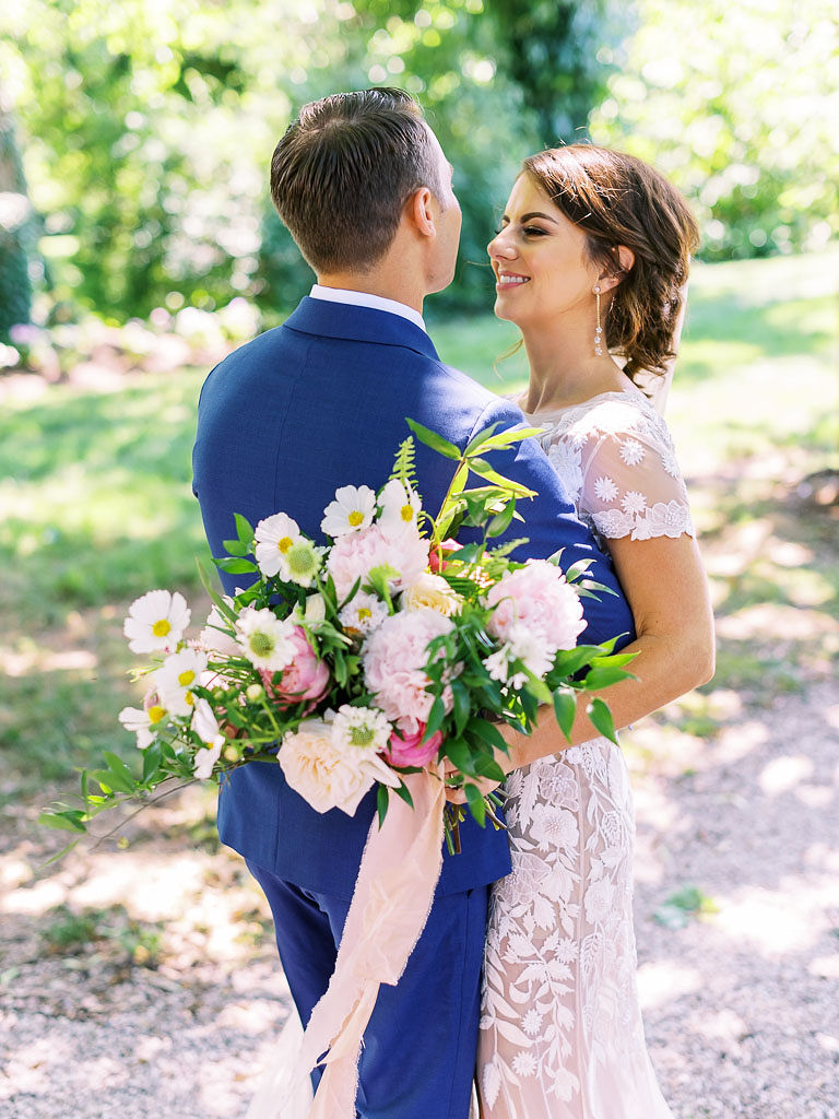 A couple embraces on their wedding day. The woman is wearing a white lace wedding gown and holding a white and pink flower bouquet. The man is wearing a deep blue suit. Taken by Virginia wedding photographer Kim Branagan.