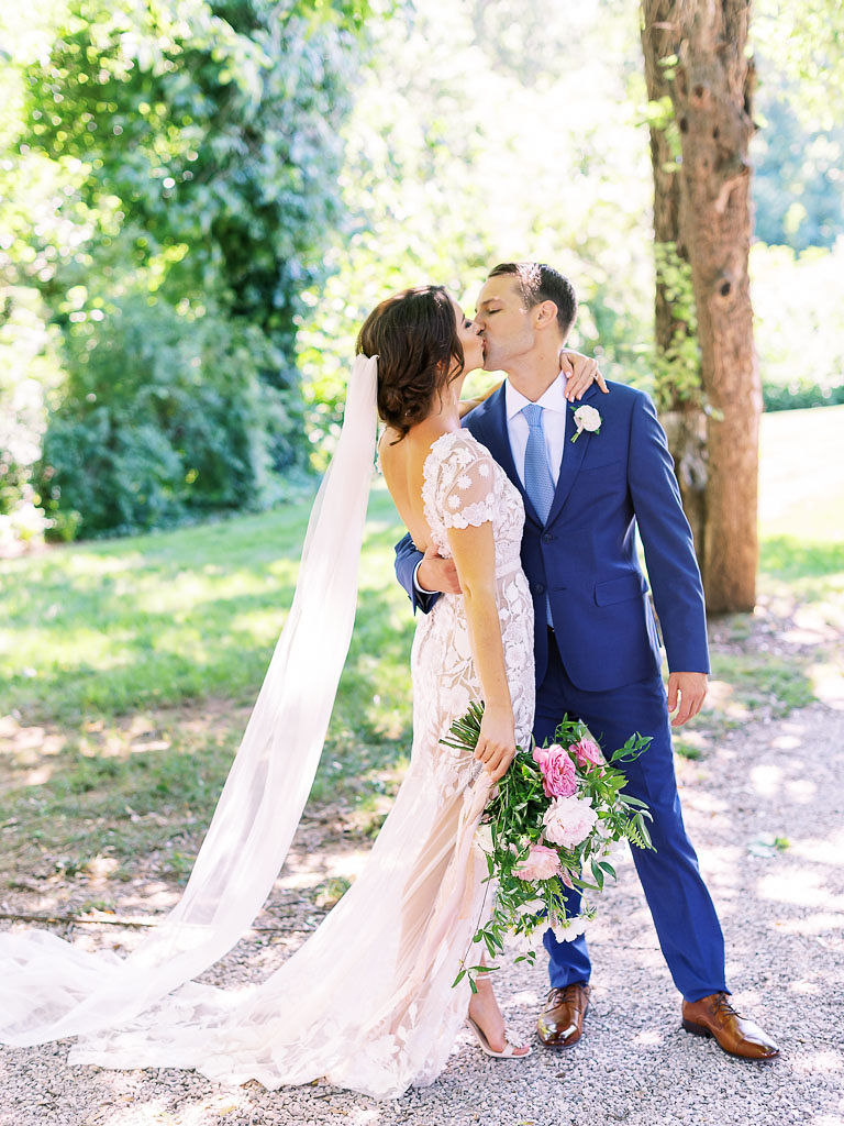 A couple embraces and kisses on their wedding day. The woman is wearing a white lace wedding gown with a long veil trailing behind her and is holding a white and pink flower bouquet. The man is wearing a deep blue suit. They are standing on a path with trees and bushes behind them. Taken by Virginia wedding photographer Kim Branagan.