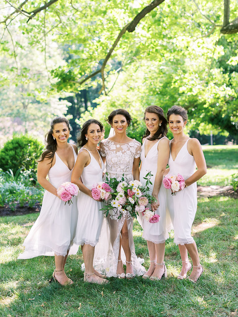 A bride wearing a white lace wedding gown standing with her bridesmaids, who are also wearing white dresses, on a green lawn with tall trees in the background. All women are holding pink and white bouquets.