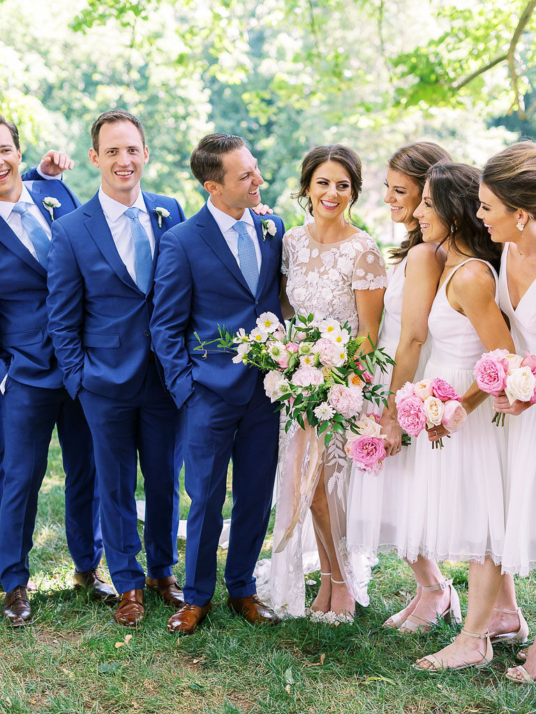 A bride and groom standing, smiling and laughing, with their bridal party. The bride is wearing a white lace wedding gown, and her bridesmaids are also wearing white dresses, on a green lawn with tall trees in the background. All women are holding pink and white bouquets. The men are all wearing matching deep blue suits.