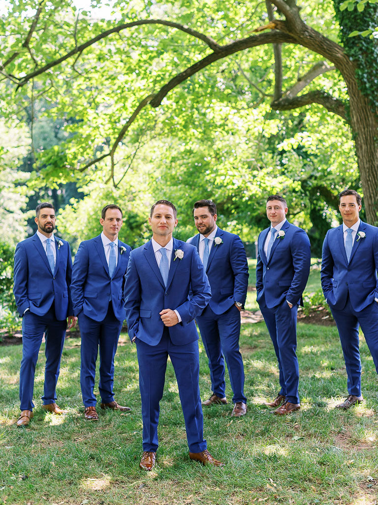 A man stands in front of his groomsmen at Antrim 1844 on his wedding day. He has his back to the groomsmen. All of men are wearing dark blue suits with blue ties.