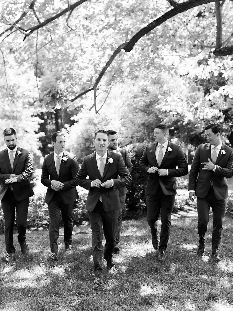 Black and white photo of a man and his groomsmen on the man's wedding day. The men are standing in a line, facing the camera, and walking together. They are all buttoning their suit coats.