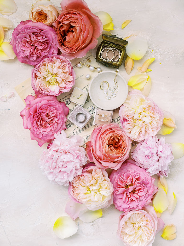 Bright florals that are various shades of pink with a couple's wedding rings and other gold jewelry arranged on a white surface.