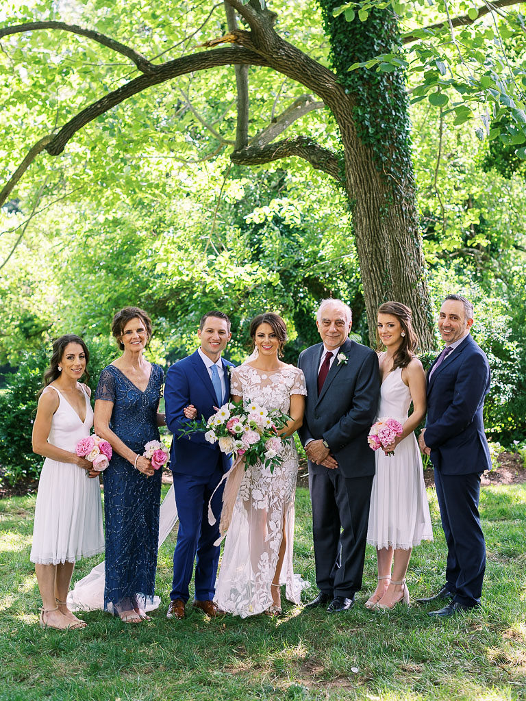 A bride, groom, and their family members standing together and smiling at the camera. The bride, her sisters, and her mother are holding pink and white bouquets. The family is all standing under a tree on green grass. Taken at Antrim 1844 by Maryland wedding photographer Kim Branagan.