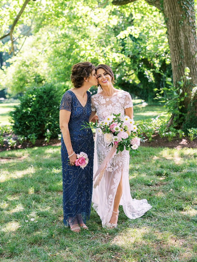 A bride and her mother stand on a green lawn with trees and bushes behind them. The bride is wearing her white lace wedding dress, and the mother is wearing a long blue dress. The mom is kissing her daughter's cheek. They are both holding pink and white floral bouquets.