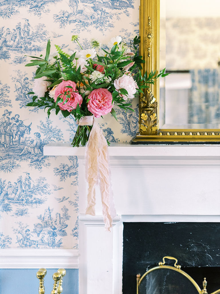 A bridal bouquet with flowers that are various shades of pink with lots of greenery sitting on a fireplace mantle, leaning against a white and blue patterned wallpapered wall. A mirror with a gold frame is also sitting on the mantle. Taken at Antrim 1844 in Maryland by D.C. and Maryland wedding photographer Kim Branagan.