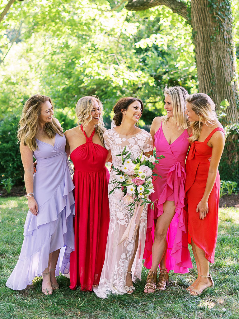 The bride stands with her friends under a large tree at Antrim 1844 in Taneytown, Maryland. They are all smiling at each other and laughing. The bride is wearing her wedding dress, and her friends are wearing lavender, pink, and red dresses.