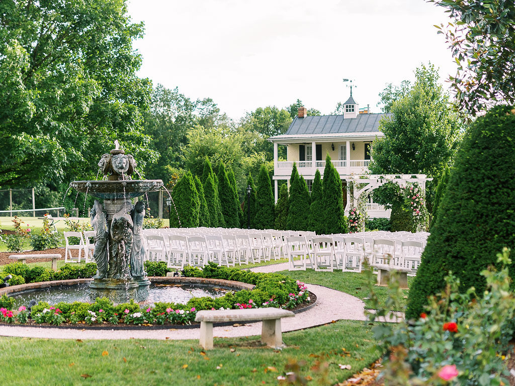 Side view of the backyard of Antrim 1844 in Taneytown, Maryland. There are white folding chairs arranged for the wedding ceremony in front of an ornate water fountain. The Antrim 1844 house is in the backyard with lots of lush, tall trees. Taken by Maryland commercial photographer Kim Branagan.