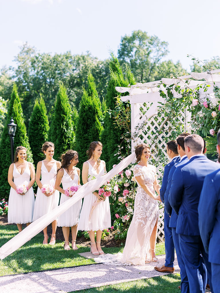 A wedding ceremony happening in front of a white trellis archway with white and pink flowers and lots of greenery hanging on the sides. The bride and groom stand in front the archway, and their bridal party stands next to them. The bride's long veil blows in the wind. Taken by Maryland, D.C., and Virginia wedding photographer Kim Branagan.