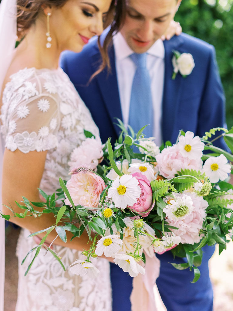 Close up shot of a bride and groom on their wedding day. The bride's dress is made of delicate lace flowers. The bride is holding a large bouquet of white and pink flowers with lots of greenery. The groom is wearing a dark blue suit, baby blue tie, and white boutonniere.