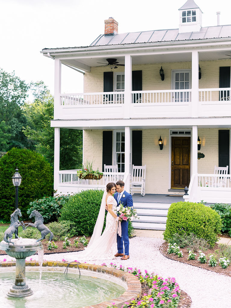 A couple embraces on their wedding day at Antrim 1844. They are standing in front of a large, cream-colored house with a porch on both the first and second stories of the home. There is an ornate water fountain in front of the couple. Taken by Maryland wedding photographer Kim Branagan.