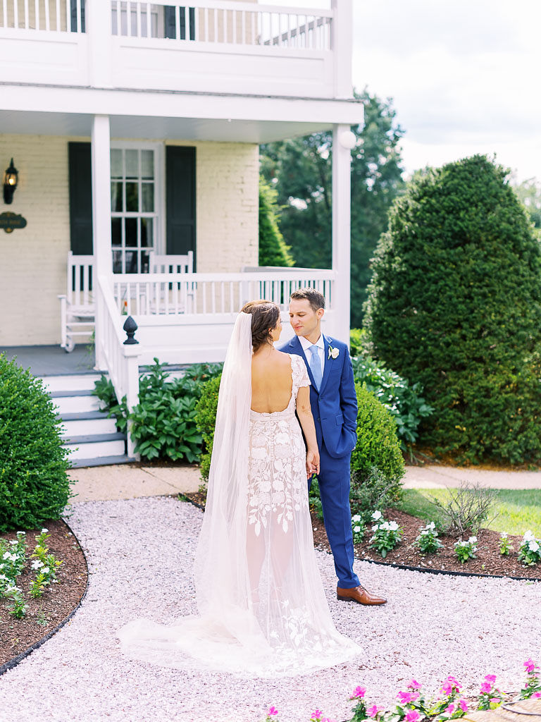 A couple faces each other and gazes into each other's eyes on their wedding day at Antrim 1844. They are standing in front of a large, cream-colored house with a porch on both the first and second stories of the home. There are many trees and shrubs surrounding the home. Taken by Maryland wedding photographer Kim Branagan.