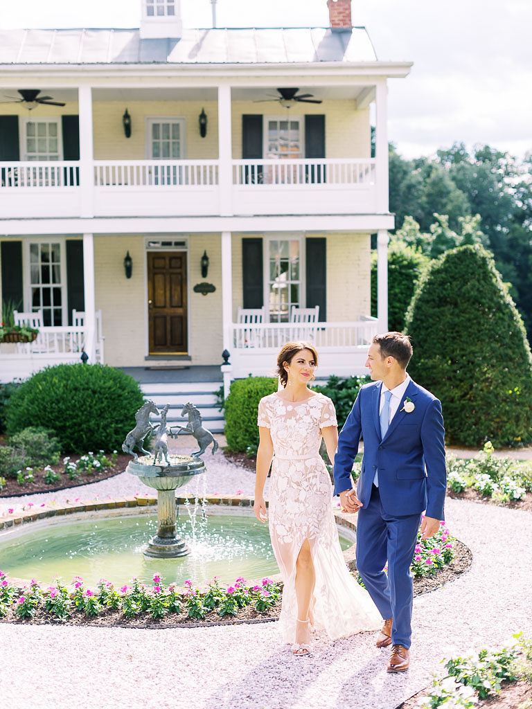 A couple holds hands and walks on a path on their wedding day at Antrim 1844. They are in front of a large, cream-colored house with a porch on both the first and second stories of the home. There is an ornate water fountain behind the couple. Taken by Maryland wedding photographer Kim Branagan.