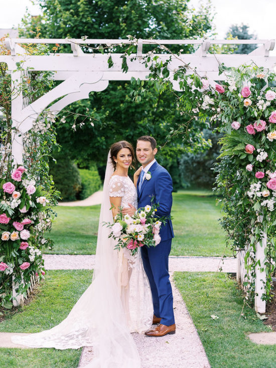 A couple stands together on their wedding day in front of a white trellis archway with white and pink flowers and lots of greenery hanging on the sides. Taken at Antrim 1844 in Taneytown, Maryland by greater DMV area wedding photographer Kim Branagan.