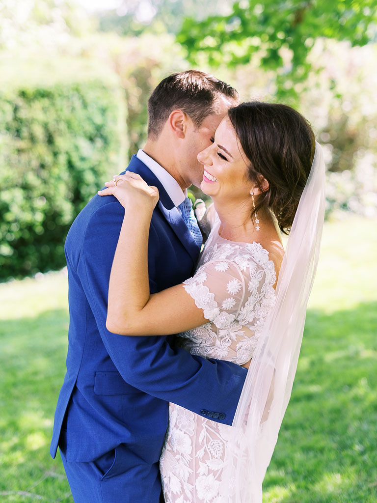 A couple embraces and laughs together standing on a green lawn on their wedding day. The bride is wearing a wedding gown made with delicate lace floral designs. The groom is wearing a dark blue suit.