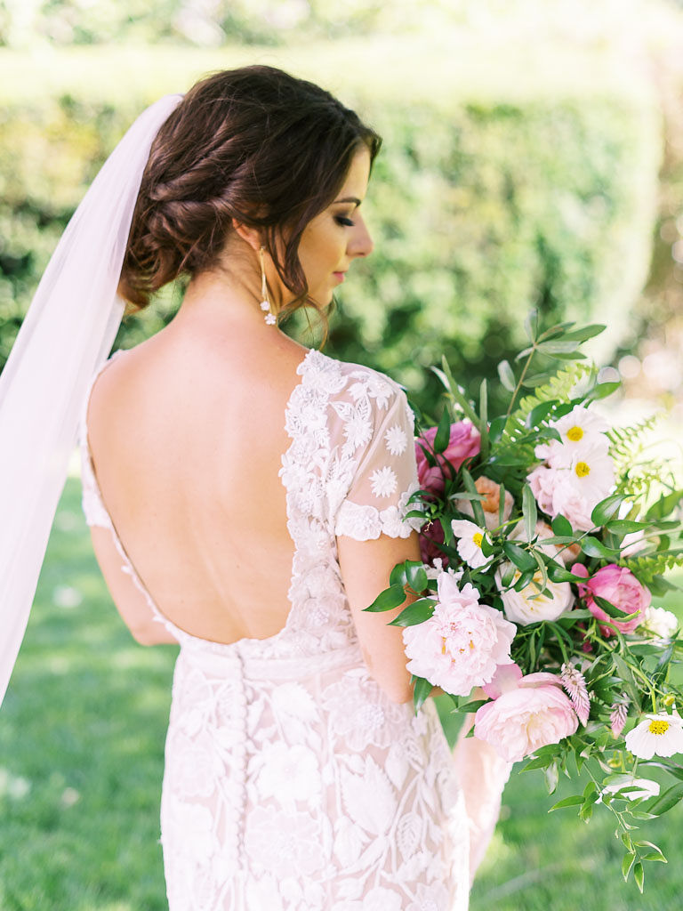 A bride stands with her back to the camera and her head turned to the side, so part of her face is shown. She is wearing her hair in a low bun and a long veil cascades down her back. Her wedding dress is made of white lace with a nude lining underneath. She is holding a large bouquet of white and pnik flowers with greenery. Taken by greater DMV area wedding photographer Kim Branagan.