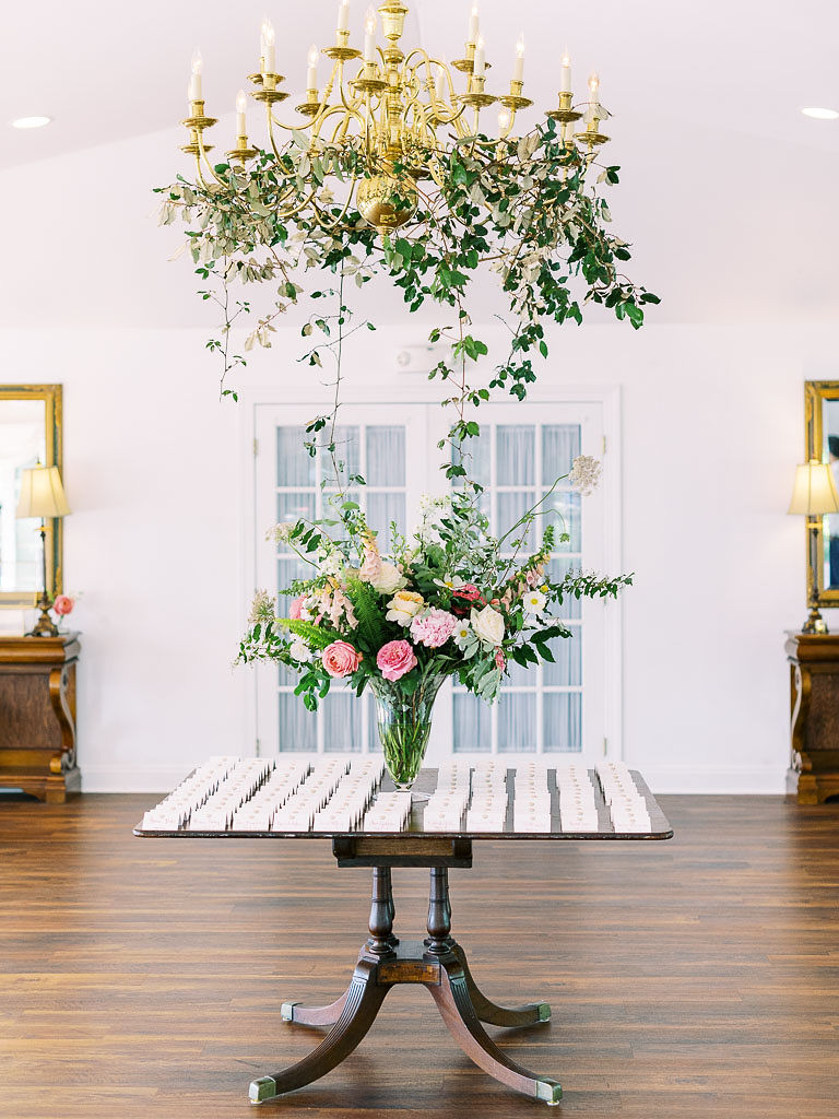 A large bouquet of white and pink blooms with lots of greenery is on a vase on a table at Antrim 1844 in Taneytown, Maryland. Above the tableis a gold chandelier with green vines hanging from it.