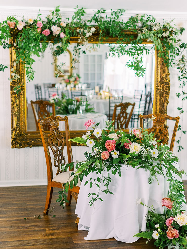 A large floral arrangement of various shades of pink flowers with pops of white flowers and tons of greenery. The florals are on a white table with two wood chairs. There is a large mirror with an ornate gold frame behind the table. There is a thick strand of green vines and some pinks flowers hanging on the top of the mirror. Taken at Antrim 1844 by Maryland wedding photographer Kim Branagan.