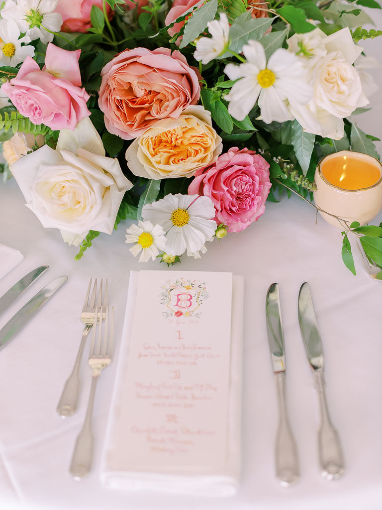 A close up shot of a place setting at a wedding reception. The table cloth is white. There is a menu, cutlery, and a white cloth napkin. The centerpiece is a lush bouquet of pink and white florals.
